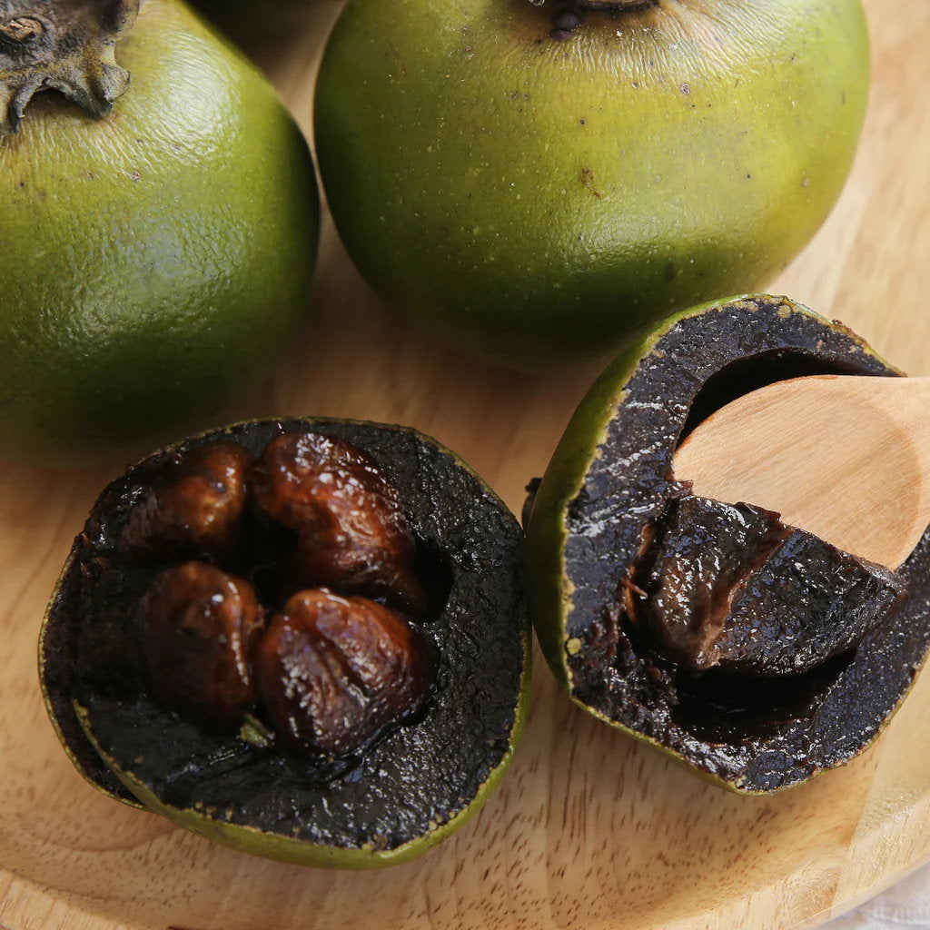 Chocolate pudding fruit, also known as black sapote, showing ripe dark brown flesh, green fruit skin, and glossy green leaves on a tropical fruit tree.